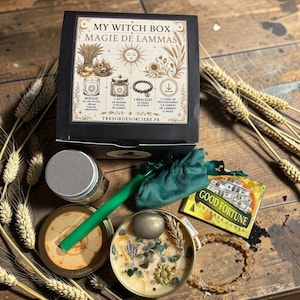 Puede incluir: Una caja negra etiquetada como My Witch Box Magie de Lammas que contiene una vela con cristales, una bolsa de tela verde, una caja de conos de incienso Good Fortune, una pulsera de cuentas, una lata de metal y un palo verde, rodeada de trigo.