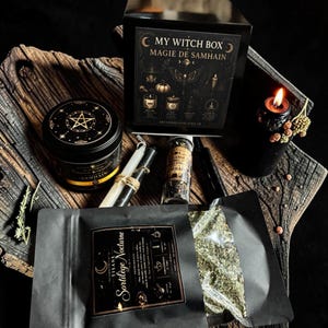 Può includere: Una scatola nera etichettata My Witch Box Magie de Samhain, un barattolo di candela nero con un pentagramma dorato, una busta nera di Tisane Sortilège Nocturne, una piccola fiala di vetro con erbe, due candele affusolate e una candela accesa.