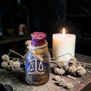 Peut inclure: Une bouteille décorative en verre avec un bouchon violet et un charme de balance argenté. La bouteille est remplie d'herbes séchées et de fleurs bleues, enveloppée de ficelle. Une bougie blanche allumée et des fleurs séchées sont en arrière-plan.