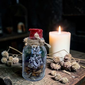 Peut inclure: Un bocal en verre transparent rempli de pierres colorées et d'un charm en forme de dragon argenté, scellé avec de la cire rouge et de la ficelle. Une bougie blanche allumée et des fleurs séchées sur une surface en bois.