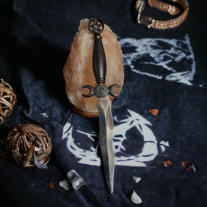 Op de afbeelding: Een zwart dolk met een pentagram en een drievoudige maanontwerp, rustend op een roze Himalaya-zoutrots. De dolk ligt op een donkerblauwe stof met witte ontwerpen.