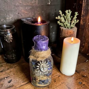 Peut inclure: Une bouteille en verre transparente avec un bouchon en cire violette et un charme soleil et lune argenté. La bouteille est enveloppée de ficelle et remplie de pierres bleues et d'herbes séchées. Deux bougies allumées et une petite plante en arrière-plan.
