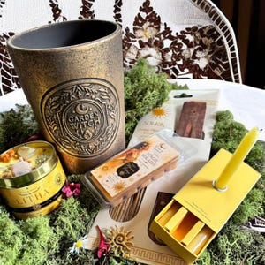 Peut inclure: Divers articles spirituels et décoratifs, dont un pot doré avec le texte "Garden Witch", une bougie jaune, une boîte de bougie "Litha", et des fondants parfumés. Les articles sont disposés sur de la mousse verte.