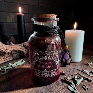 Puede incluir: Un frasco de vidrio transparente con una poción de baño de color rojo oscuro, cerrado con un tapón de corcho. La etiqueta dice "Ancient Witch Seduction Bath Spell" y "240g". Un cordón negro rodea el cuello con un colgante de cristal morado.