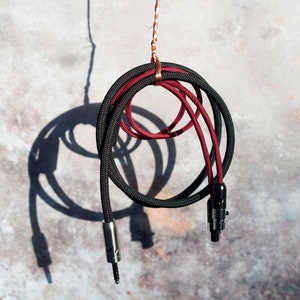 Może przedstawiać: Czarny i bordowy kabel audio ze srebrnymi i czarnymi złączami. Kabel jest zapętlony i zawieszony na nakrapianym szarym tle, rzucając cień. Kabel jest przeznaczony do sprzętu audio.