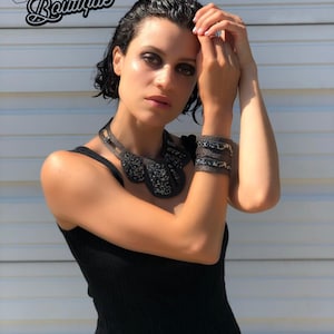 May include: A woman wearing a black tank top with a beaded necklace and a black beaded bracelet. The background is a white wall with horizontal lines.