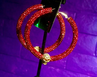 May include: Red rhinestone hoop earrings with a gold skull charm. The earrings are displayed on a black stand against a purple background. The text "JONIQUÉ Boutique" is visible in the bottom right corner of the image.