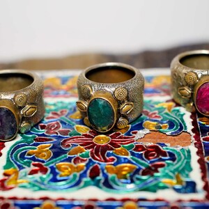 Könnte beinhalten: Drei verzierte Ringe mit großen ovalen Edelsteinen. Die Ringe haben ein silberfarbenes Band mit goldfarbenen Akzenten. Die Edelsteine sind lila, grün und rot. Die Ringe werden auf einer farbenfrohen, gemusterten Oberfläche präsentiert.