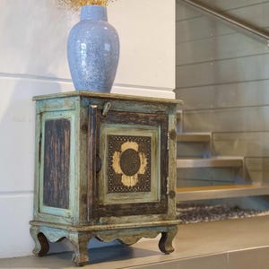 May include: A small, antique wooden cabinet with a distressed finish in shades of teal, brown, and gold. The cabinet has a decorative front panel with a central design and is topped with a blue vase.