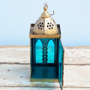 May include: A brass lantern with a teal glass panel and intricate cutouts. The lantern has a domed top with a hanging loop.