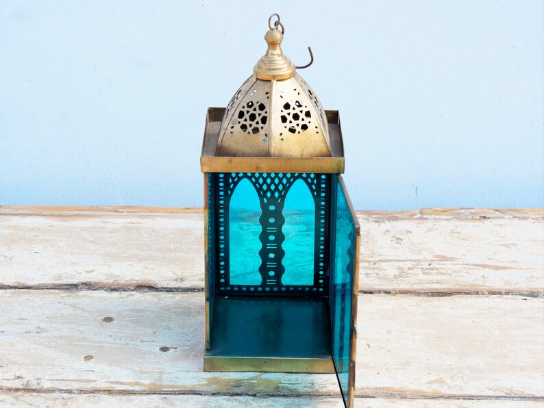 May include: A brass lantern with a teal glass panel and intricate cutouts. The lantern has a domed top with a hanging loop.