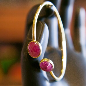 Può includere: Un bracciale color oro con due pietre preziose ovali rosso intenso. Il bracciale ha un design curvo e aperto, con una gemma nella parte superiore e un'altra nella parte inferiore. Le gemme sono incastonate in castoni dorati.