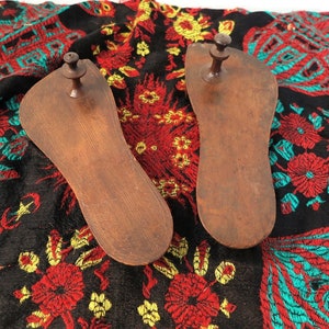 Può includere: Due forme per scarpe in legno scuro, ciascuna con un piccolo piolo rotondo. Sono posizionate su un tessuto nero con un vivace motivo floreale in rosso, giallo e turchese. L'intricato design del tessuto crea uno sfondo colorato.