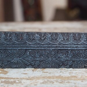 Puede incluir: Un bloque rectangular negro con un diseño tallado. El diseño presenta un patrón repetitivo de hojas y flores.