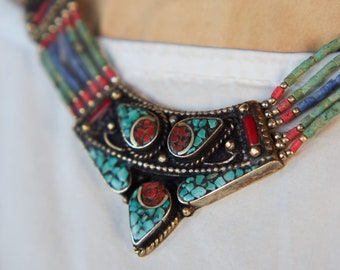 May include: Close-up of a handcrafted necklace featuring turquoise, red, and blue beads. The necklace has a central pendant with turquoise and red stone inlays, set in a dark metal frame with gold accents. The necklace is worn on a white shirt.