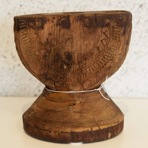 Può includere: Oggetto decorativo in legno fatto a mano con finitura marrone scuro. L'oggetto ha una parte superiore arrotondata con motivi geometrici intagliati e una base svasata. Un filo bianco è legato al centro.