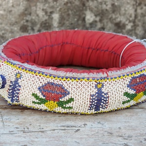 Puede incluir: Un collar de cuentas con una base de tela roja y cuentas blancas, azules, amarillas y rojas formando un patrón floral.