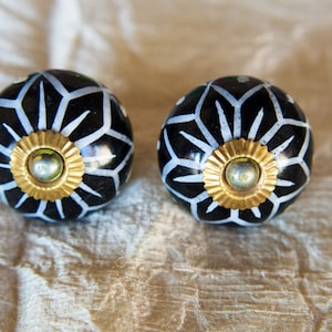 May include: Two black ceramic cabinet knobs with white geometric designs and gold metal accents.