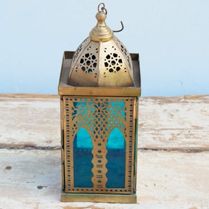 May include: A gold metal lantern with intricate cutouts and a blue glass panel. The lantern has a domed top and a square base.