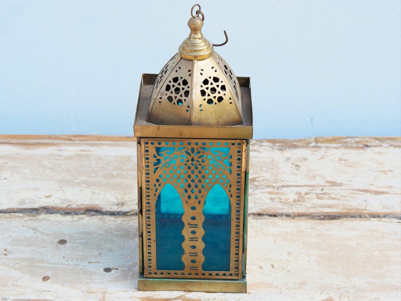 May include: A gold metal lantern with intricate cutouts and a blue glass panel. The lantern has a domed top and a square base.