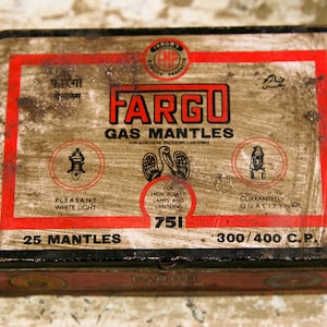 Może przedstawiać: Prostokątna metalowa puszka z Fargo Gas Mantles do lamp ciśnieniowych na naftę. Puszka jest brązowa z czerwonym obramowaniem i tekstem. Tekst zawiera "FARGO" dużymi czerwonymi literami, a także "25 Mantles" i "300/400 C.P."
