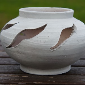 Può includere: Vaso in ceramica bianca con disegni ritagliati. Il vaso ha una forma arrotondata e un'ampia apertura.