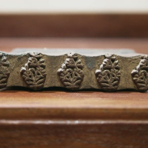 Op de afbeelding: Een close-up van een houten drukblok met vier ingewikkelde bloemmotieven die in het oppervlak zijn gegraveerd. Het blok is bruin en heeft een licht verweerde uitstraling.