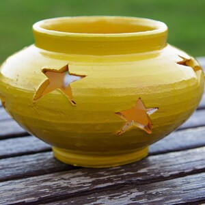 Può includere: Un portacandele in ceramica giallo con tagli a forma di stella. Il portacandele è su una superficie di legno.