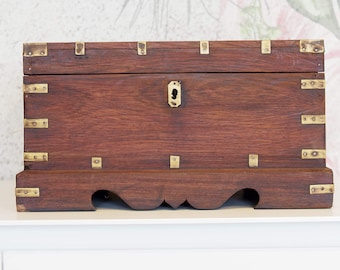 May include: A rectangular wooden chest with a dark brown finish and brass corner brackets and accents. The chest has a keyhole and sits on a decorative base. The chest is likely used for storage.