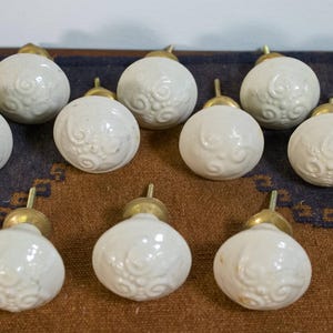 May include: Ten white ceramic cabinet knobs with a decorative floral design. The knobs have a gold metal base.