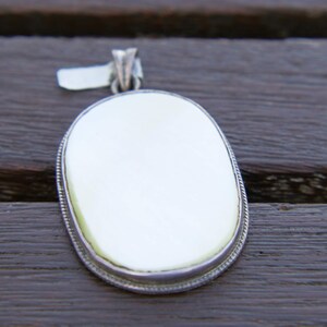 Może przedstawiać: Owalny wisiorek z jasnym, białym środkiem i srebrną, sznurową obwódką. Wisiorek ma małą pętlę u góry na łańcuszek. Tło to ciemna drewniana powierzchnia.