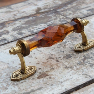 Puede incluir: Un tirador de puerta de cristal facetado de color ámbar con herrajes de latón. El tirador tiene un diseño geométrico curvo y está montado sobre placas de latón decorativas. La ferretería tiene una estética vintage.