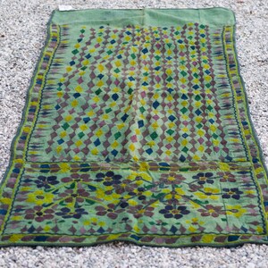 May include: A green fabric rug with a geometric pattern of diamonds in shades of green, yellow, blue, and pink. The rug has a border of floral designs in shades of green, yellow, and brown.