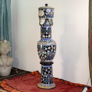 Peut inclure: Un vase en céramique décoratif haut avec un fond noir et des motifs floraux et géométriques colorés. Le vase a un couvercle avec le texte "ROYAL CAFE & CIGARE" en lettres noires.