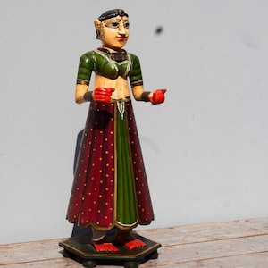Puede incluir: Una figurita de madera de una mujer que lleva una falda roja y verde con detalles dorados. Lleva una blusa verde y manos rojas. La figurita está de pie sobre una base de madera.