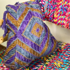 May include: A purple, blue and gold embroidered cushion with a geometric pattern. The cushion is sitting on a colourful woven rug.