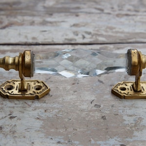 Peut inclure: Une poignée de porte en verre clair et facetté avec des ferrures en laiton. Les composants en laiton comprennent des plaques de base hexagonales décoratives et des embouts cylindriques. La poignée en verre présente un motif en losange.