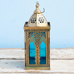 May include: A gold metal lantern with intricate cutouts and a blue glass panel. The lantern has a traditional Moroccan design.