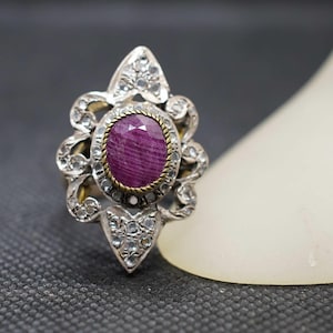 Puede incluir: Anillo de plata ornamentado con una gran gema de rubí ovalada. El rubí está rodeado por un bisel dorado y acentuado con gemas claras más pequeñas. El anillo tiene un diseño detallado de inspiración floral.