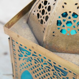 May include: A gold metal lantern with intricate cut-out designs and a blue glass panel. The lantern has a traditional Moroccan style.