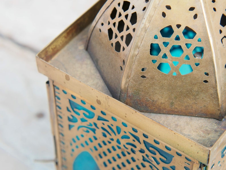 May include: A gold metal lantern with intricate cut-out designs and a blue glass panel. The lantern has a traditional Moroccan style.