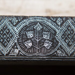 May include: A close-up of a black metal printing block featuring an intricate design of a floral motif and geometric patterns. The design is carved into the surface of the block, creating a raised effect.