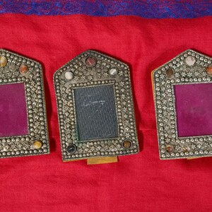 Puede incluir: Tres marcos de fotos ornamentados, de color plateado, con una abertura rectangular y un borde decorativo de pequeñas piedras y adornos. Los marcos tienen un respaldo morado y están sobre un fondo rojo.