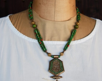 May include: A multi-strand necklace with green and red beads and a large, ornate pendant. The pendant is a hexagonal shape with intricate metalwork and a green stone center.