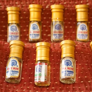 Op de afbeelding: Een verzameling kleine, heldere parfumflesjes met gele doppen, gerangschikt op een rode, getextureerde ondergrond. Elk flesje heeft een etiket met tekst en een decoratief embleem. De flesjes bevatten verschillende geuren, waaronder "Blue Musk" en "White Musk".