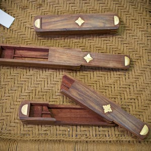 Puede incluir: Tres cajas de madera con detalles de latón. Las cajas son rectangulares con tapa corredera. Cada caja tiene un elemento decorativo dorado en el centro. Las cajas se exhiben sobre una superficie tejida.