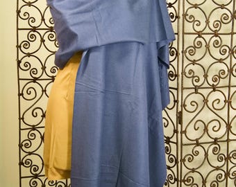 May include: A blue shawl draped over a mannequin. The shawl is a solid color and has a fringed edge. The mannequin is wearing a mustard-colored garment. A decorative metal screen is in the background.