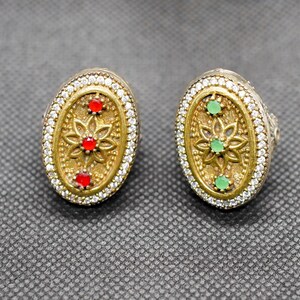 May include: Two ornate, oval-shaped rings. Each ring features a gold-colored central design with a floral pattern and small red or green gemstones. The outer edges are lined with small, clear gemstones, creating a sparkling effect. The rings are set against a textured gray background.