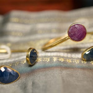 Può includere: Bracciali a polsino aperti color oro con accenti di pietre preziose. Un bracciale presenta una gemma rotonda rosa intenso, mentre altri mostrano pietre blu a forma di goccia. I gioielli poggiano su un tessuto grigio fantasia.