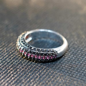 May include: A silver ring with a band of black and pink gemstones. The ring is set against a dark, textured background. The gemstones are small and closely set, creating a sparkling effect. The ring is a piece of jewelry.